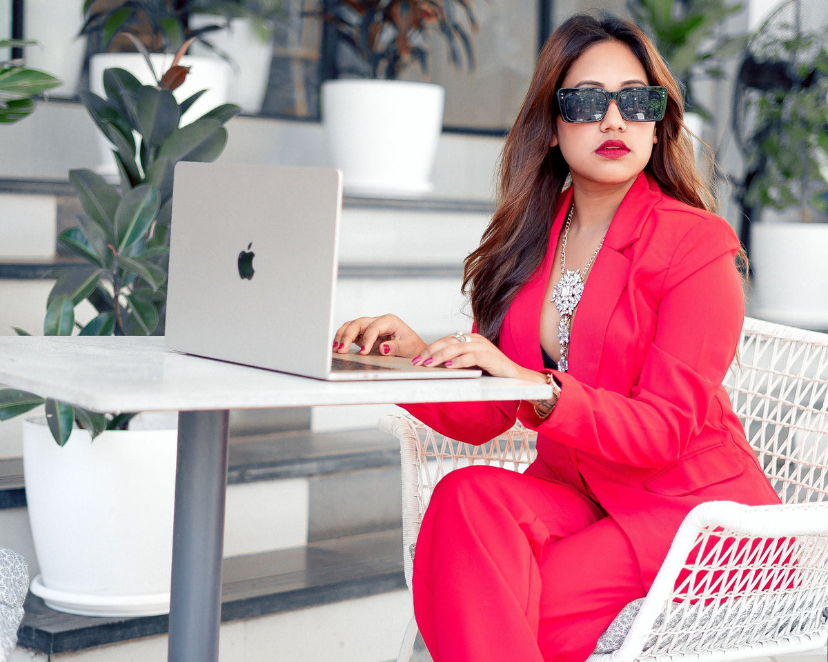 A woman wearing a bright red two-piece suit and sunglasses sits at a white table with a laptop in a modern office lobby with potted plants.